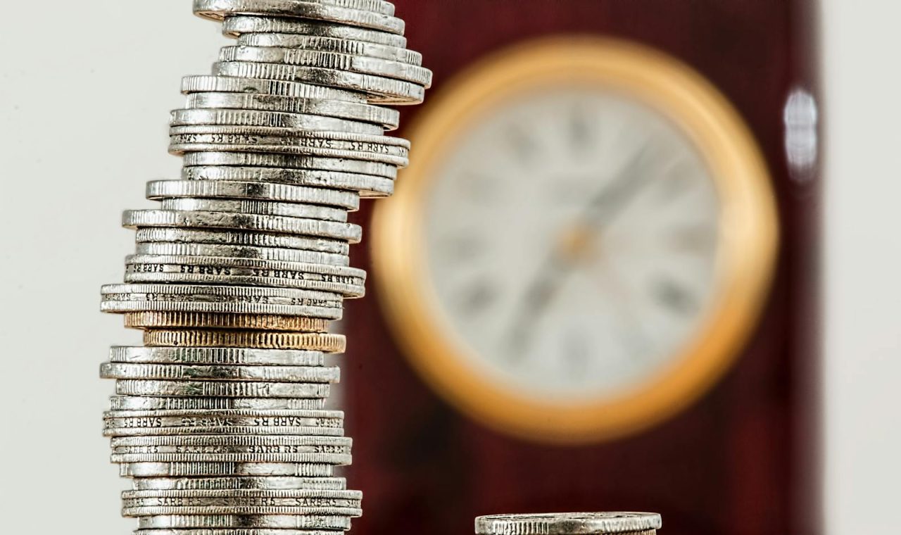 A close-up image of stacked coins with a blurred clock, symbolising time and money relationship