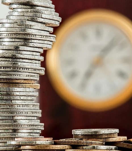 A close-up image of stacked coins with a blurred clock, symbolising time and money relationship