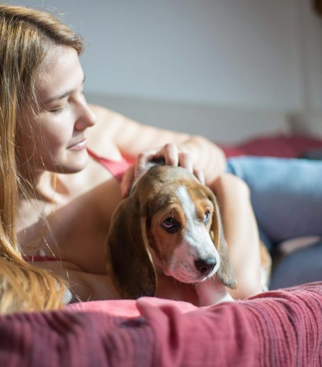 A young woman lies on a couch, embracing her beagle dog in a warm indoor setting